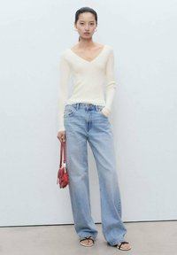 Crème v-halstrui, lichtblauwe wijde jeans en zwarte flip-flops. Accessoires omvatten een rode fringetas. Soepele textuur en casual pasvorm.