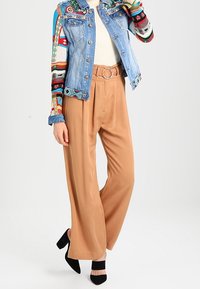 Nainen, jolla on yllään vaaleanruskeat leveälahkeiset housut, joissa on renkaallinen vyö, värikkäällä kuviolla varustettu pitkähihaisen paidan, farkkutakin ja mustat korkokengät.