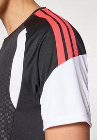 Αθλητική μπλούζα με κοντό μανίκι που διαθέτει μαύρο σώμα, λευκές και κόκκινες λεπτομέρειες, υφή με μοτίβο και τρεις κόκκινες ρίγες στον ώμο.