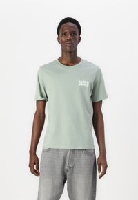Mintgrønn bomull t-skjorte med rund hals og korte ermer. Har hvit logo trykt på venstre bryst. Båret med lysegrå denimjeans.
