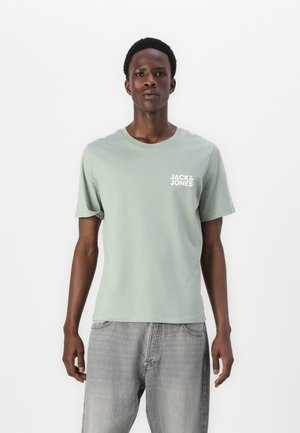 Mint green cotton t-shirt with a crew neck and short sleeves. Features white logo print on the left chest. Worn with light gray denim jeans.