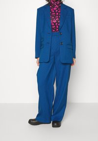 Completo a quadri blu con giacca strutturata, pantaloni a gamba larga e dolcevita nera con motivo floreale rosa. Calzature nere.