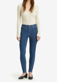 Mujer vistiendo un cárdigan de botón de canalé en crema, jeans skinny de tiro alto en azul con doble cierre de botón, y zapatos de tacón bajo negros.