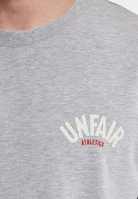 Szara bawełniana koszulka z okrągłym dekoltem, z napisem "UNFAIR" w białym kolorze i "ATHLETICS" w czerwonym, delikatnie teksturowana tkanina.