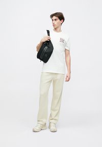 T-shirt branca com estampa preta, calças de treino bege e ténis castanhos. Bolsa tiracolo preta com detalhe de fecho. Fundo liso.