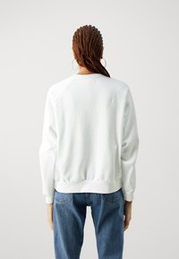 Felpa bianca con maniche raglan, polsini e orlo a coste, texture morbida, abbinata a jeans denim blu. Vista da dietro, finitura liscia.