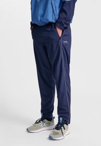 Navy trackbyxor med elastisk midja och sidofickor, med kontrasterande grå piping. Tillsammans med grå och blå sportskor.