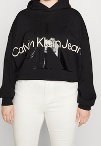 Czarna krótka bluza z kapturem z dużym, błyszczącym logo Calvin Klein. Posiada sznurek w kapturze i długie rękawy. W zestawieniu z białymi dżinsami.