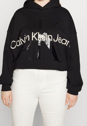 Haut court noir à capuche avec un grand logo brillant Calvin Klein. Présente une capuche avec cordon de serrage et des manches longues. Associé à un jean blanc.