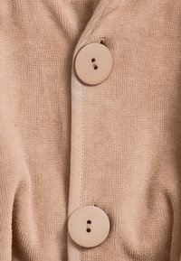 Ljust beige tyg med en strukturerad yta, med två stora, runda knappar i matchande färg längs en central söm.