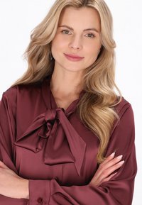 Bordó színű szatén blúz masnival a gallérján, hosszú ujjakkal és gombos mandzsettával. Simaságú anyag enyhe fényes hatással.