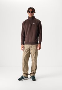 Brown fleece quarter-zip pullover, beige cargo pants with pockets, and green athletic shoes with black accents. Circular sunglasses add detail.
