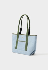 Světle modrá taška na rameno se zelenými madly a lemem, se přední kapsou s malým zlatým knoflíkem, stojící na bílém pozadí.