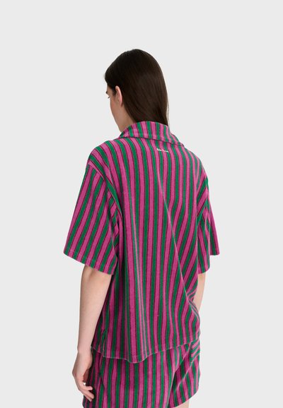 Personne aux longs cheveux foncés portant un haut ample à manches courtes et un short avec des rayures verticales vertes, roses et noires, vue de dos sur fond blanc.