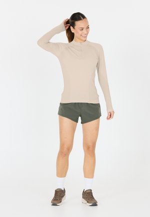 Beige langärmliges Sportoberteil mit einem Reißverschluss am Hals, kombiniert mit olivgrünen Shorts. Das Model trägt weiße Socken und braune Sneakers.