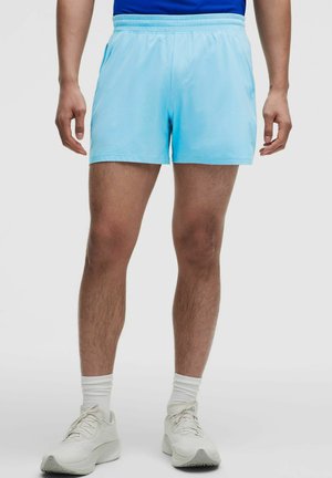 Homme portant un short de sport bleu clair, des chaussettes blanches et des chaussures de course blanches, debout sur un fond uni.