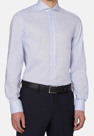 Muž nosící světle modrou košili s dlouhým rukávem zastrčenou do černých kalhot s černým pásem, stojící s rukama uvolněně po stranách.