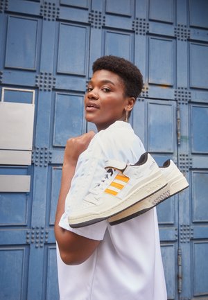 Person i hvid skjorte bærer hvide sneakers med orange striber over skulderen og står foran en blå dør med paneler.
