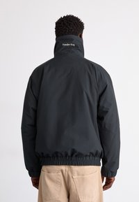 Schwarze Nylonjacke mit hohem Kragen, elastischen Bündchen und Saum; mit "Powder Keg"-Logo auf dem Rücken, glatter Textur und lockerer Passform.