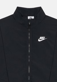 Črna Nike jakna z visokim ovratnikom, sprednjim zadrgo in belim logotipom. Tkanina se zdi gladka in lahka. Ni vidnih vzorcev ali okrasov.