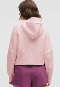 Rosa, kurz geschnittenes Hoodie mit einer Kordelzugkapuze, das aus strukturiertem Stoff besteht und über gewellte Nahtdetails verfügt, kombiniert mit lila Sportshorts.