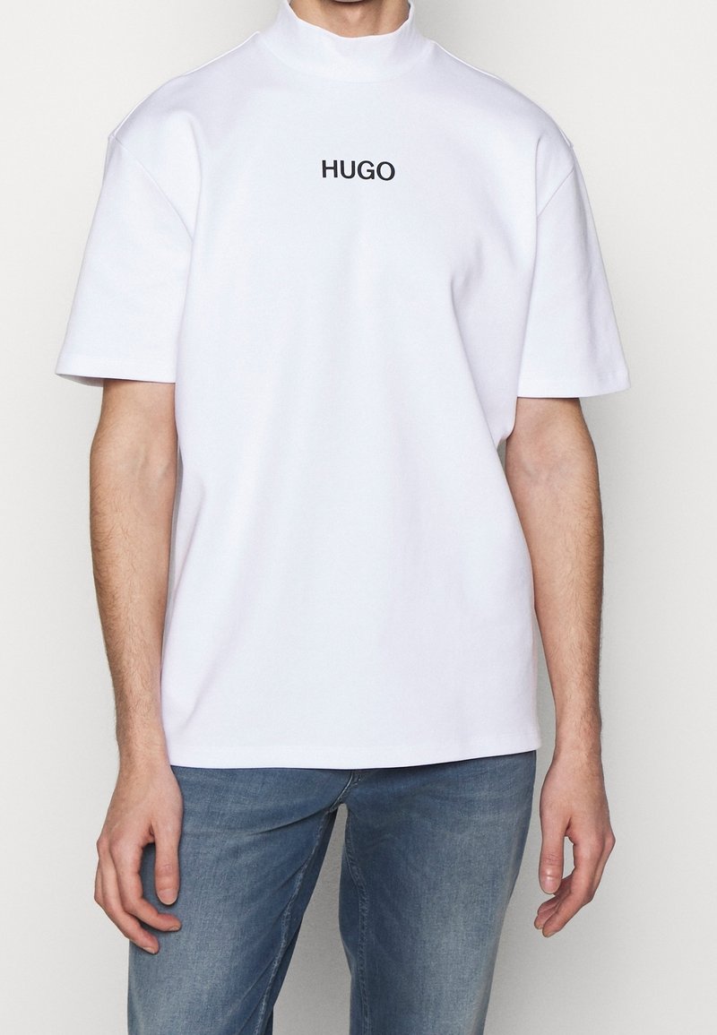 T-shirt blanc à col montant et manches courtes avec le texte "HUGO" sur la poitrine, porté par une personne en jean bleu, sur fond blanc uni.