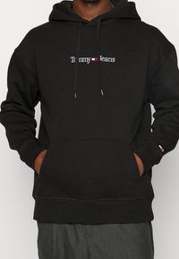 Sudadera negra con el texto y logo de "Tommy Jeans" en el pecho, manos en el bolsillo delantero, usada con pantalones grises.