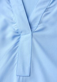 Blouse bleu clair avec un col en V et des détails froncés. Tissu doux et lisse avec une couture propre autour du col et une texture subtile.