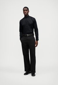 Camisa preta de manga comprida com gola mandarim, combinada com calças pretas de perna direita e sapatos pretos brilhantes, apresentando um design elegante.