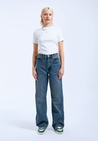 Witte kortekous T-shirt gecombineerd met high-waisted blauwe denim wijde pijpen jeans en groene accent sneakers, met een relaxte pasvorm.