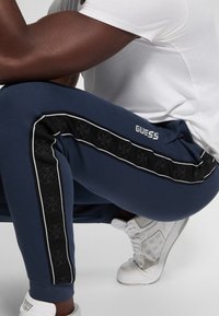 Pantaloni jogger blu navy con strisce laterali nere caratterizzati da un motivo logo, piping bianco e logo "GUESS". Abbinati a sneakers bianche.