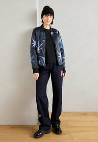 Blouson noir avec des motifs abstraits bleus, t-shirt noir, jeans en denim foncé avec broderies florales, et chaussures noires épaisses.