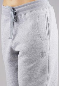 Waist and upper thigh of person wearing light gray sweatpants with side pocket, drawstring tie, and subtle hexagon logo on thigh.