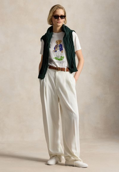 Femme portant des lunettes de soleil, un t-shirt blanc avec un imprimé d'ours en peluche, un pantalon blanc à jambes larges, une ceinture marron, des baskets blanches et un pull vert foncé posé sur les épaules.