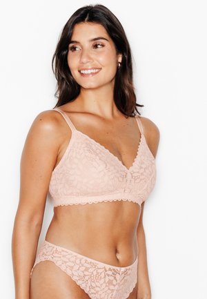 Femme aux cheveux foncés portant un soutien-gorge en dentelle rose clair assorti et une culotte, souriante et regardant vers sa gauche sur fond blanc.