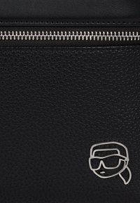 Nahaufnahme von schwarzem strukturiertem Leder mit silbernem Reißverschluss und kleinem silbernem Logo in Umrissform eines stilisierten Gesichts mit Sonnenbrille und Kopftuch.