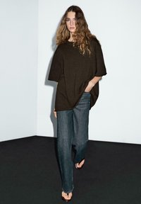 Maglione morbidamente oversize di colore marrone scuro con maniche corte, abbinato a jeans di denim scuro a vestibilità ampia e infradito marroni. Sfondo minimalista.