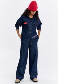 Jeansowy strój składający się z koszuli zapinanej na guziki z kieszenią na piersi i haftowanymi detalami, zestawiony z szerokimi spodniami, oba w ciemnym niebieskim kolorze z kontrastującymi czerwonymi akcentami.