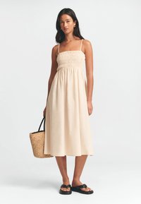 Cremefarvet midi-kjole med smock-buster og tynde stropper, der har en flydende nederdel. Parret med sorte sandaler og en vævet håndtaske.