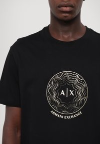 T-shirt en coton noir avec un logo doré circulaire portant le texte "A/X" et "Armani Exchange", entouré de motifs de points décoratifs.