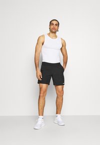 Weißes ärmelloses Tanktop und schwarze Shorts mit Nike-Logo, kombiniert mit weißen Sneakers. Einfaches Design, lässige Passform und sportlicher Stil.