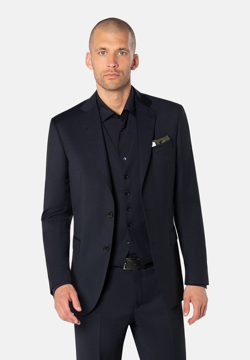 Mann mit rasiertem Kopf, der einen dunkelblauen Dreiteiler mit schwarzem Hemd und Einstecktuch trägt, steht vor einem schlichten hellen Hintergrund.