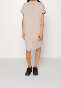 Robe chemise beige à manches courtes avec un col et une fermeture à boutons, associée à des sabots noirs. Texture lisse, longueur au genou.