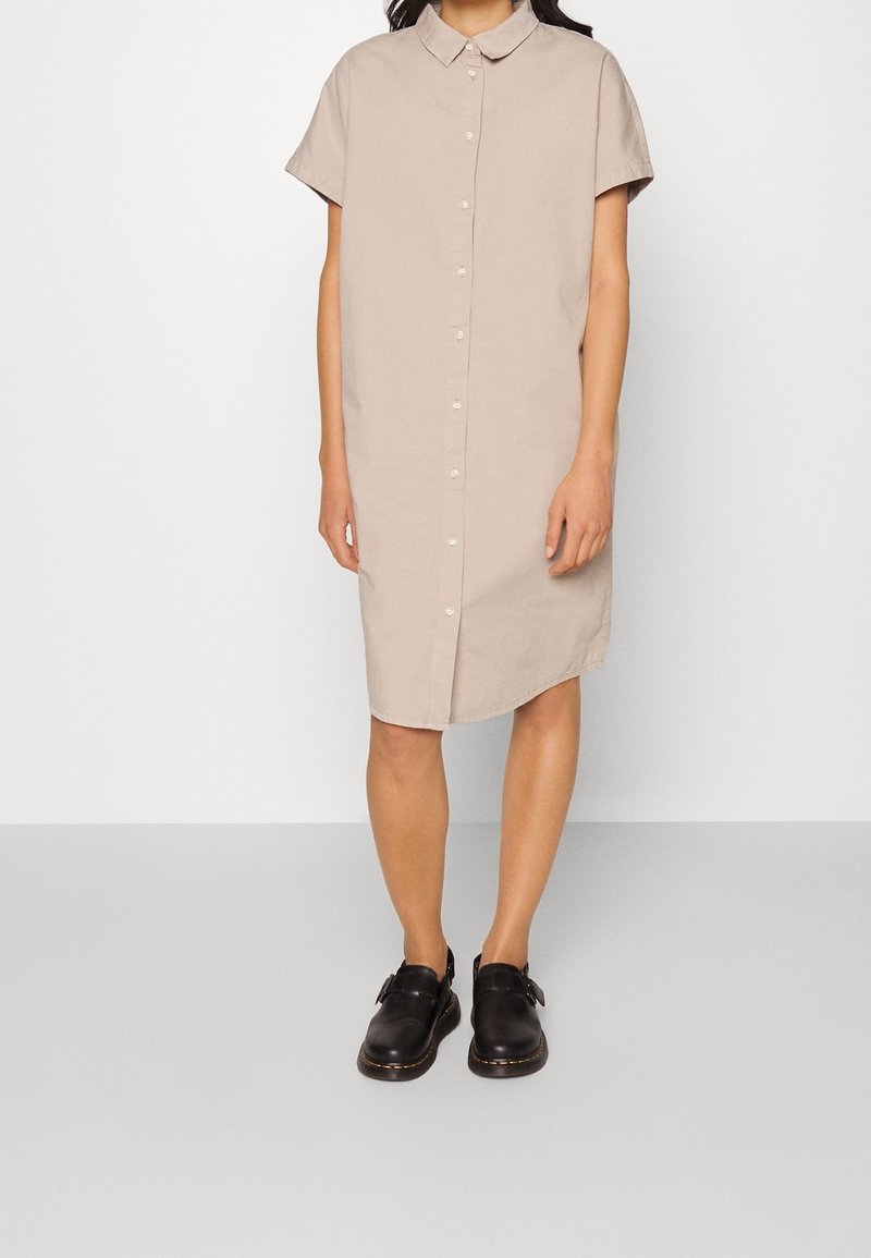Robe chemise beige à manches courtes avec un col et une fermeture à boutons, associée à des sabots noirs. Texture lisse, longueur au genou.