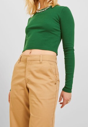 Femme portant un crop top vert à manches longues, un pantalon beige taille haute et un collier chaîne, visible du milieu de la poitrine jusqu'aux cuisses.