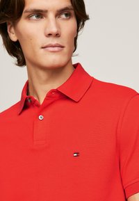 Röd polo t-shirt i texturerad tyg, med krage och en liten logotyp broderad på vänster bröst.