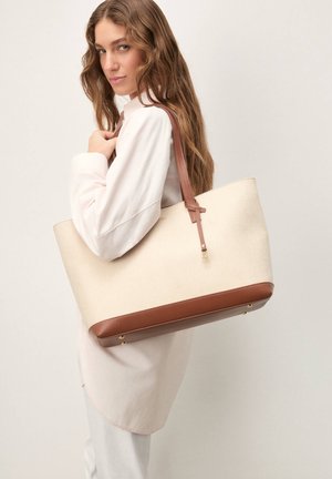 Ženska z valovitimi lasmi, oblečena v svetla oblačila, nosi veliko bež torbo za na ramo z rjavimi usnjenimi ročaji in obrobami.