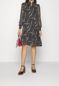 Vestido negro con patrón ondulado en beige, mangas largas con puños fruncidos, escote tipo ojal y dobladillo fluido en capas. Se lleva con tacones gruesos negros.