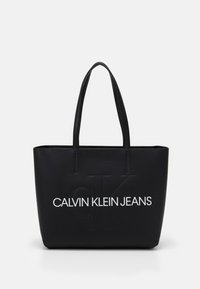 Svart tote-väska gjord av slätt syntetmaterial, med präglad logotyp "CALVIN KLEIN JEANS" i vitt, dubbla robusta handtag, rektangulär form.