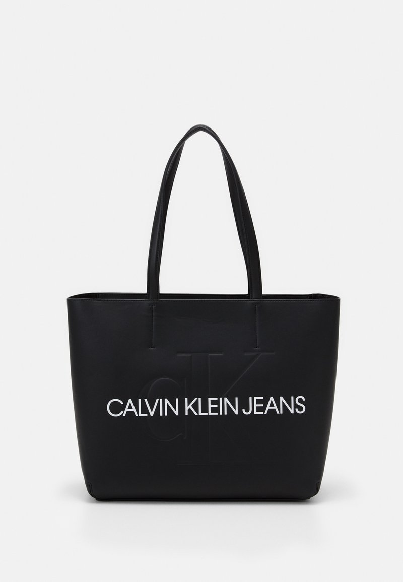 Svart tote-väska gjord av slätt syntetmaterial, med präglad logotyp "CALVIN KLEIN JEANS" i vitt, dubbla robusta handtag, rektangulär form.